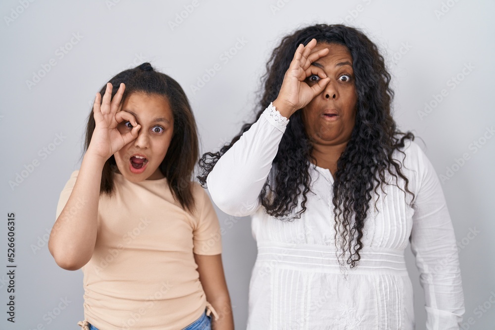 Mother and young daughter standing over white background doing ok gesture shocked with surprised face, eye looking through fingers. unbelieving expression.