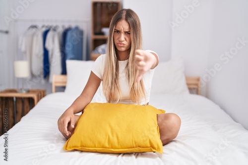 Young blonde woman sitting on the bed with pillow at home looking unhappy and angry showing rejection and negative with thumbs down gesture. bad expression.