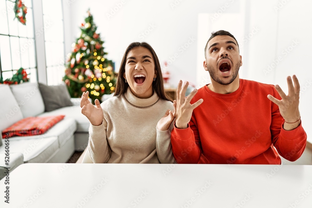 Fototapeta premium Young hispanic couple sitting on the table by christmas tree crazy and mad shouting and yelling with aggressive expression and arms raised. frustration concept.