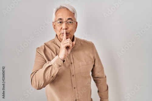 Hispanic senior man wearing glasses asking to be quiet with finger on lips. silence and secret concept.