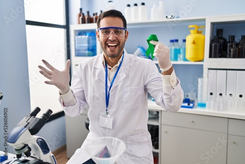 Young hispanic man with beard working at scientist laboratory holding green ribbon celebrating victory with happy smile and winner expression with raised hands