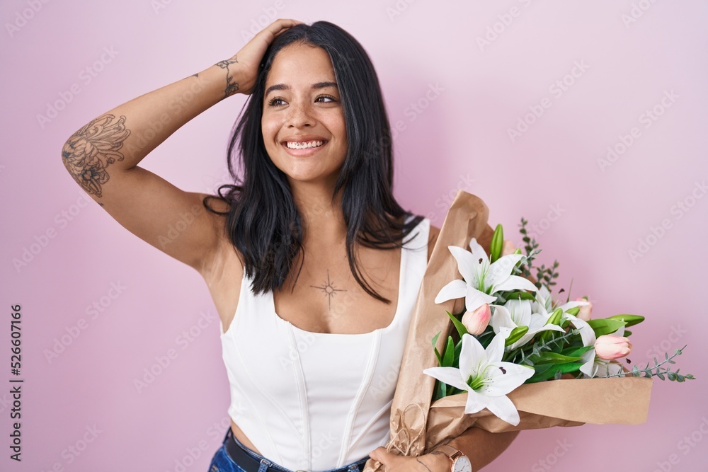 Brunette woman holding bouquet of white flowers smiling confident touching hair with hand up gesture, posing attractive and fashionable
