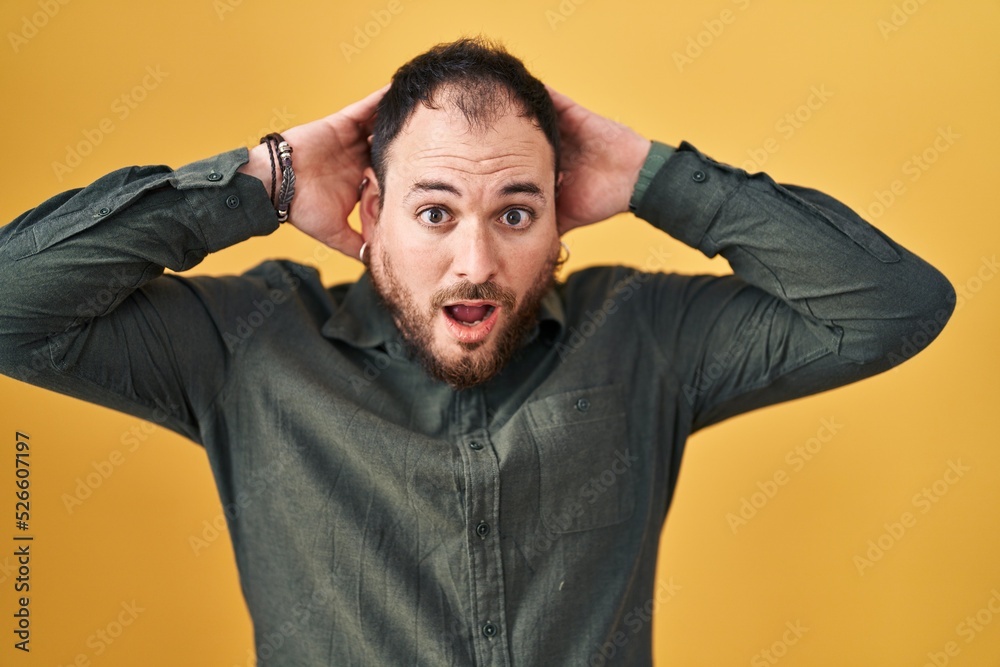 Plus size hispanic man with beard standing over yellow background crazy and scared with hands on head, afraid and surprised of shock with open mouth Plus size hispanic man with beard standing over yellow background crazy and scared with hands on head, afraid and surprised of shock with open mouth