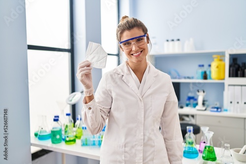 Fototapeta Naklejka Na Ścianę i Meble -  Young blonde woman wearing scientist uniform holding medical mask at laboratory
