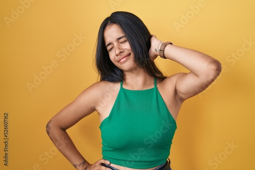 Brunette woman standing over yellow background suffering of neck ache injury, touching neck with hand, muscular pain