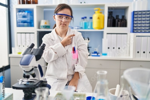 Hispanic girl with down syndrome working at scientist laboratory pointing with hand finger to the side showing advertisement  serious and calm face