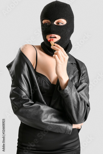 Young woman in balaclava applying red lipstick against light background