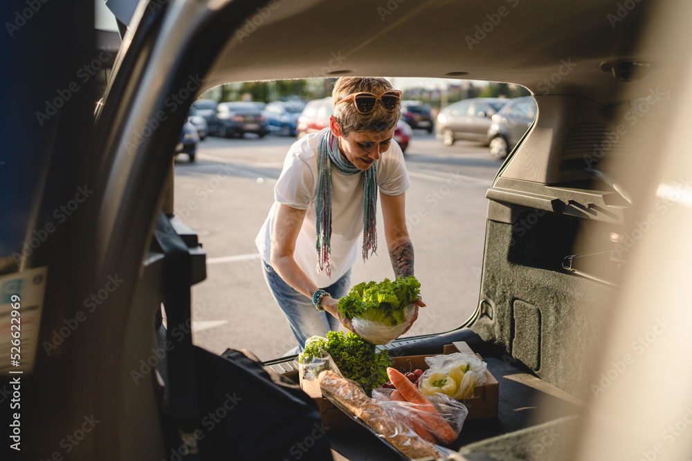 One woman mature caucasian female putting box with vegetables in the back of her car in sunny day on the parking lot in front of supermarket or grocery store in sunny day copy space real people
