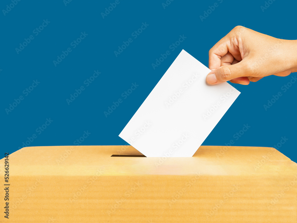 Hand voter holding ballot paper putting into the voting box at place election against a blue background Hand voter holding ballot paper putting into the voting box at place election against a blue background