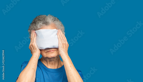 Senior woman using hot compress gel on eyes while standing on a blue background photo