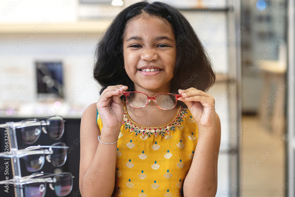 Happy Indian little girl is choosing a new glasses in optician store