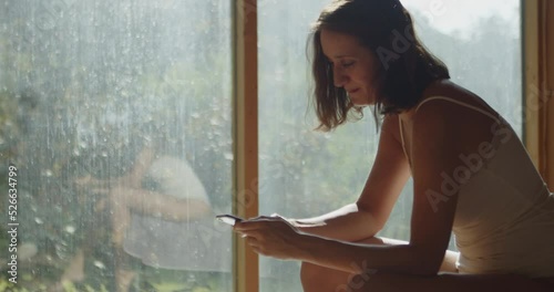 A young half-naked woman sitting on a bed in the morning sun, texting on her cell phone