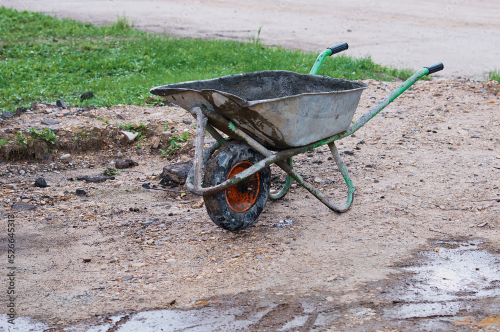An old dirty metal wheelbarrow with an orange wheel. Used construction tool, side view. Agricultural Wheel Farm Cart Flat Lawns Garden Cart Wheel Hand Cart An old dirty metal wheelbarrow with an orange wheel. Used construction tool, side view. Agricultural Wheel Farm Cart Flat Lawns Garden Cart Wheel Hand Cart