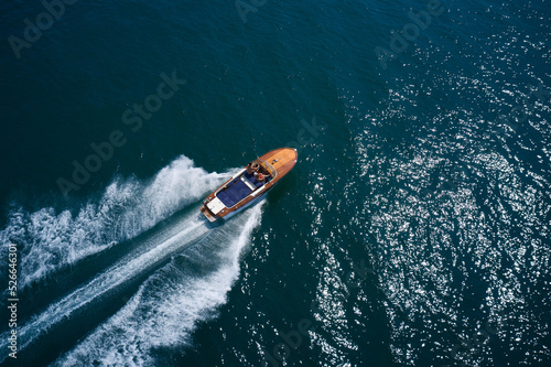 An expensive wooden boat is an average movement on the water. Classic wooden boat with motor moving on blue water aerial view. Italian wooden boat fast moving diagonal top view.