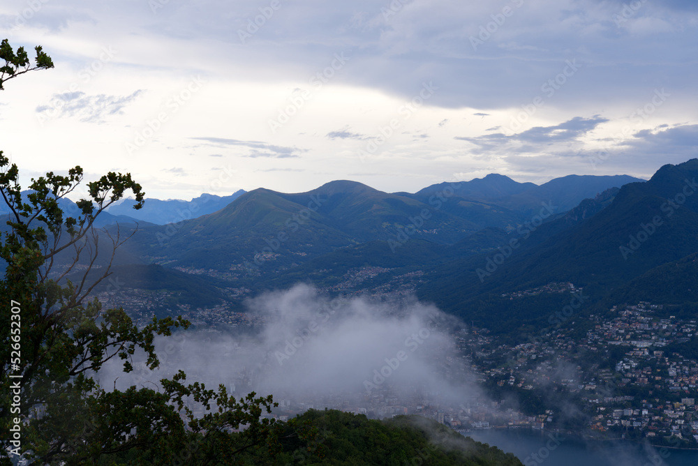 Obraz premium Scenic landscape with mountain panorama and Lake Lugano seen from local mountain San Salvatore at City of Lugano on a cloudy summer day. Photo taken July 4th, 2022, Lugano, Switzerland.