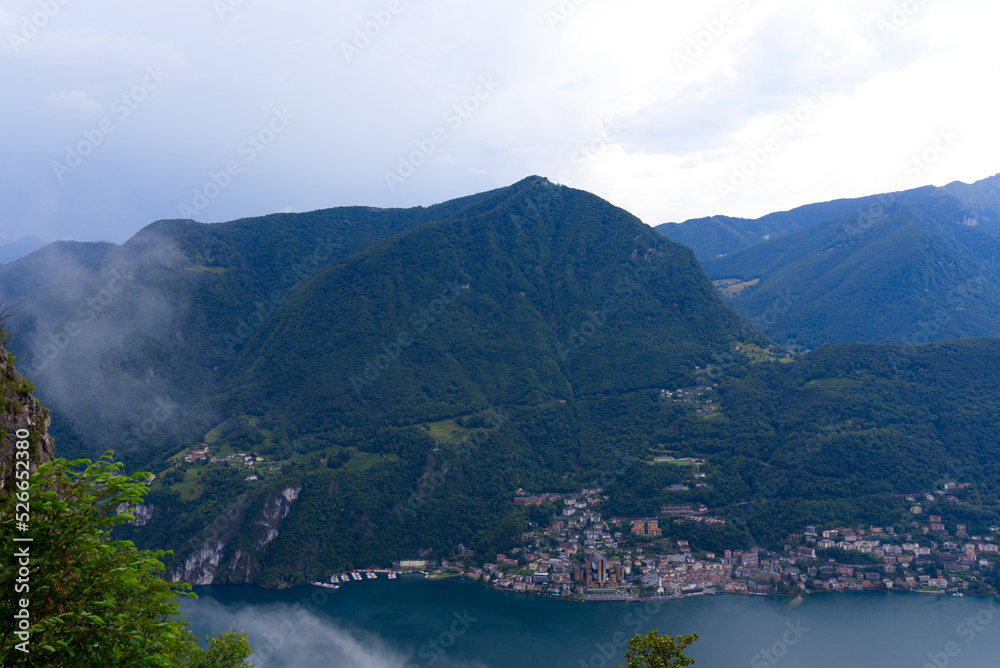 Aerial view of mountains and lake with Italian Village of Campione seen from local mountain San Salvatore at City of Lugano on a cloudy summer day. Photo taken July 4th, 2022, Lugano, Switzerland.