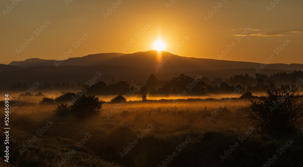 Early morning scenery in field and mountains. Sun casting beautiful rays of light through the mist and trees.