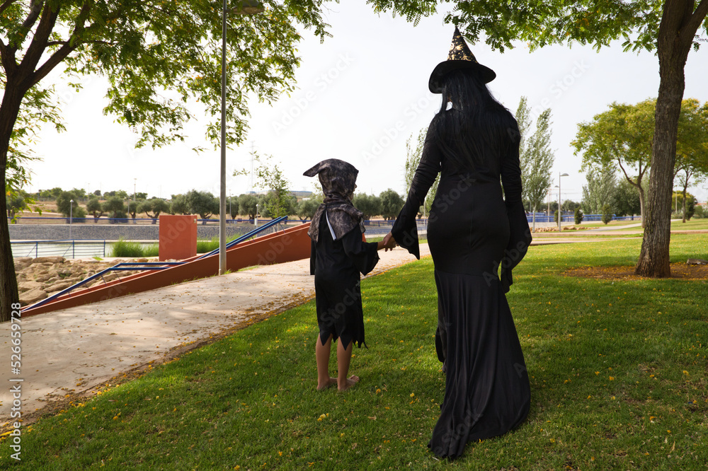Obraz premium Happy halloween. Child dressed up for halloween party and his mother dressed as a witch walking through the park seen from behind. Trick or treat. 31st October