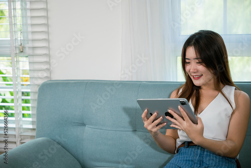 Happy Asian woman playing smart tablet at home while relaxing on sofa living room, Beautiful female sitting on couch using digital tablet pc, technology social media