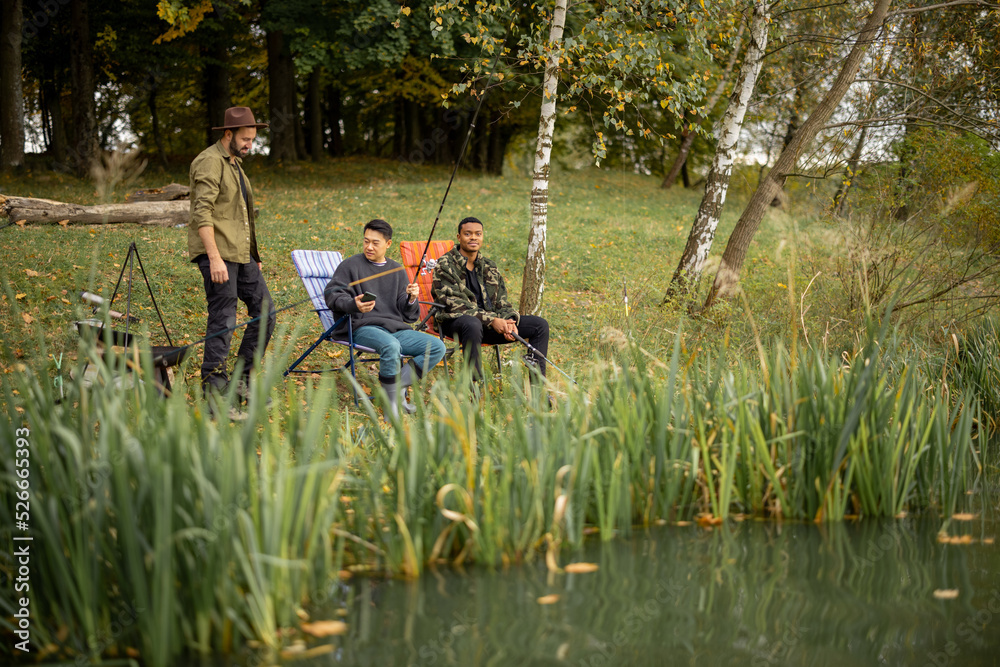 Pleased multiracial male friends resting and fishing on river or lake coast. Concept of leisure, hobby and weekend in nature. Idea of friendship and spending time together. View from the waterside Pleased multiracial male friends resting and fishing on river or lake coast. Concept of leisure, hobby and weekend in nature. Idea of friendship and spending time together. View from the waterside