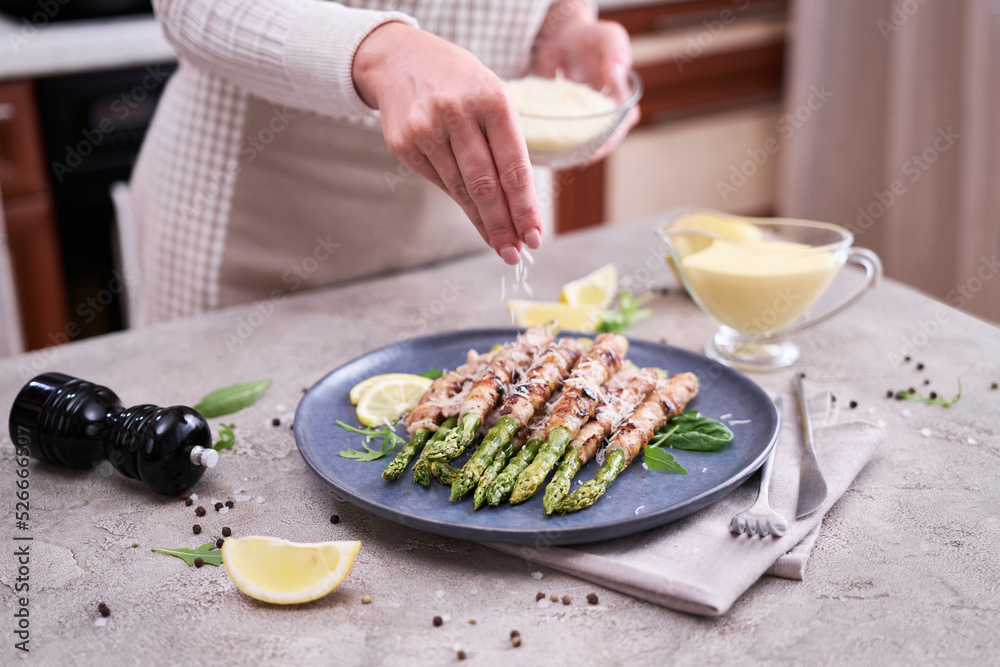 woman pouring grated parmesan cheese on a Asparagus wrapped with bacon and spices on a plate