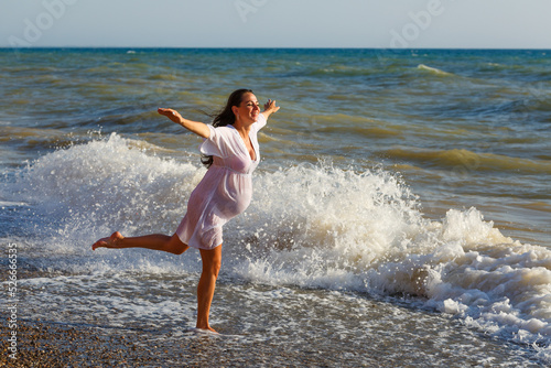 Pregnant woman is jumping on beach. Cheerful pregnant woman runs on seashore. Pregnant woman in a romantic white dress jumping on the beach near the tropical sea. Indication of happy pregnant women.