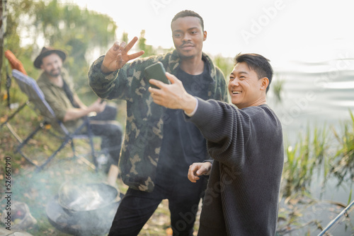Smiling multiracial male friends taking selfie on smartphone during resting in nature. Men fishing and cooking Ukha soup on river coast at autumn. Leisure, weekend and vacation. Idea of friendship © rh2010