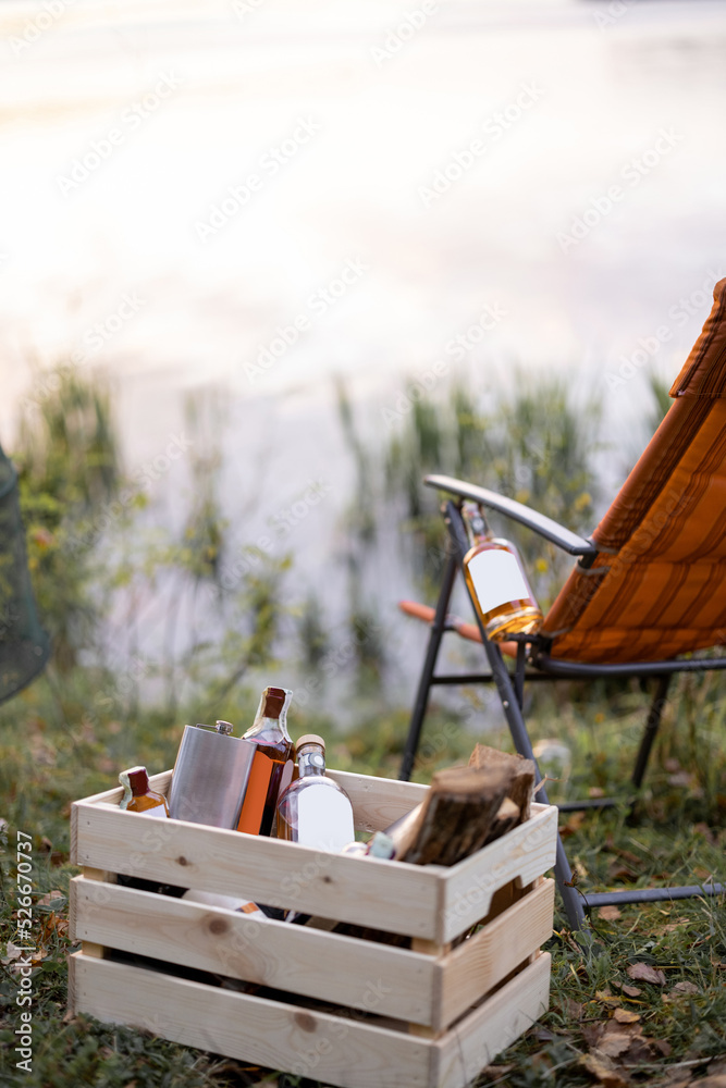 Obraz premium Bottles with strong alcohol at wooden box and folding chair at picnic or fishing. Bottle with blank label to copy paste. Mockup image for branding drink