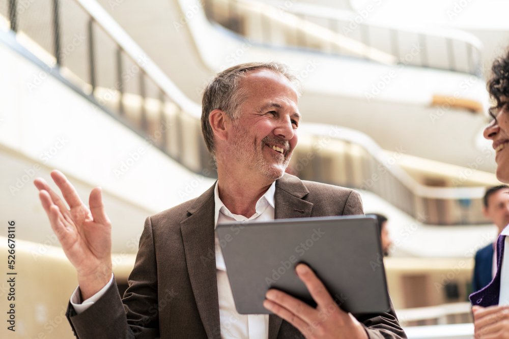Smiling businessman holding tablet PC discussing with colleague in corridor Smiling businessman holding tablet PC discussing with colleague in corridor