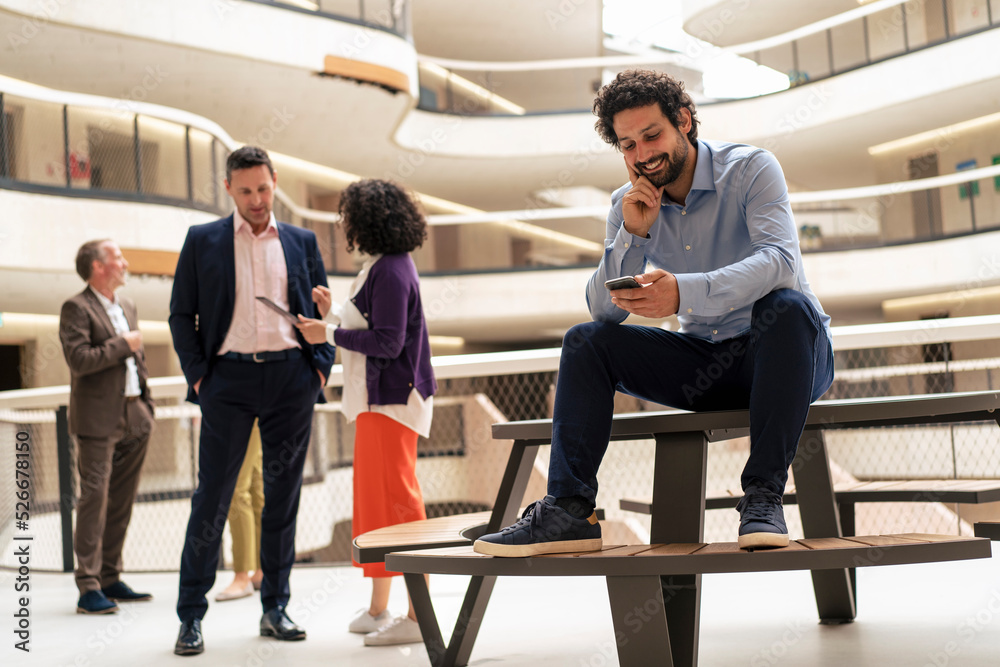 Smiling businessman using smart phone with colleagues discussing in background at corridor Smiling businessman using smart phone with colleagues discussing in background at corridor