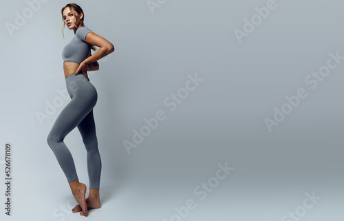 Fototapeta Naklejka Na Ścianę i Meble -  Beautiful young woman with natural make-up and gathered hair, dressed in a gray sports uniform, posing in the studio on a gray background.Advertising sportswear and yoga wear. Healthy lifestyle, sport