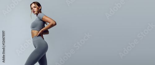 Beautiful young woman with natural make-up and gathered hair, dressed in a gray sports uniform, posing in the studio on a gray background. Advertising sportswear and yoga wear. 
