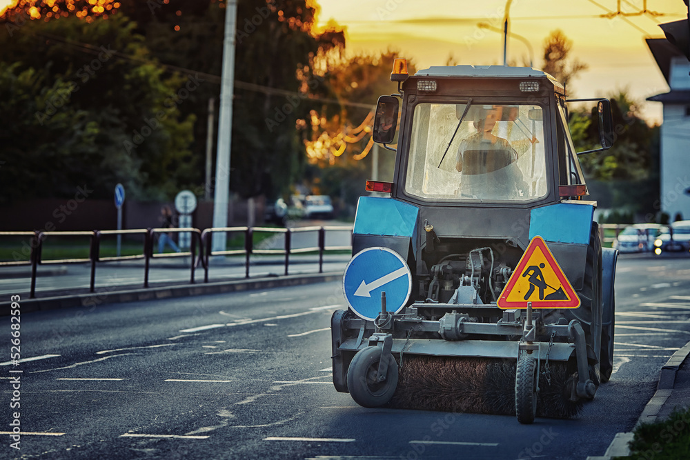 Obraz premium Tractor clean street. Tractor with rotating brush sweep road from dust and dirt. Municipal street cleaning vehicle. City cleaning machine on the city road.