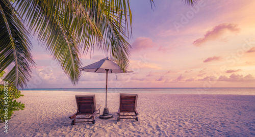Beautiful tropical island sunset, couple sun beds chairs umbrella under palm tree leaves. White sand sea view horizon idyllic evening sky. Inspire beach resort panorama. Romantic paradise beach banner