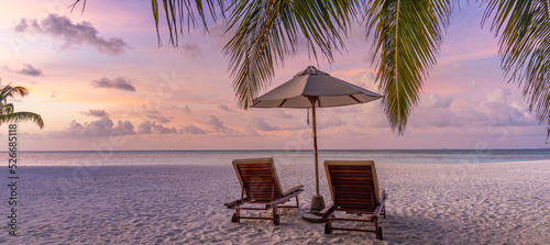 Beautiful tropical island sunset, couple sun beds chairs umbrella under palm tree leaves. White sand sea view horizon idyllic evening sky. Inspire beach resort panorama. Romantic paradise beach banner