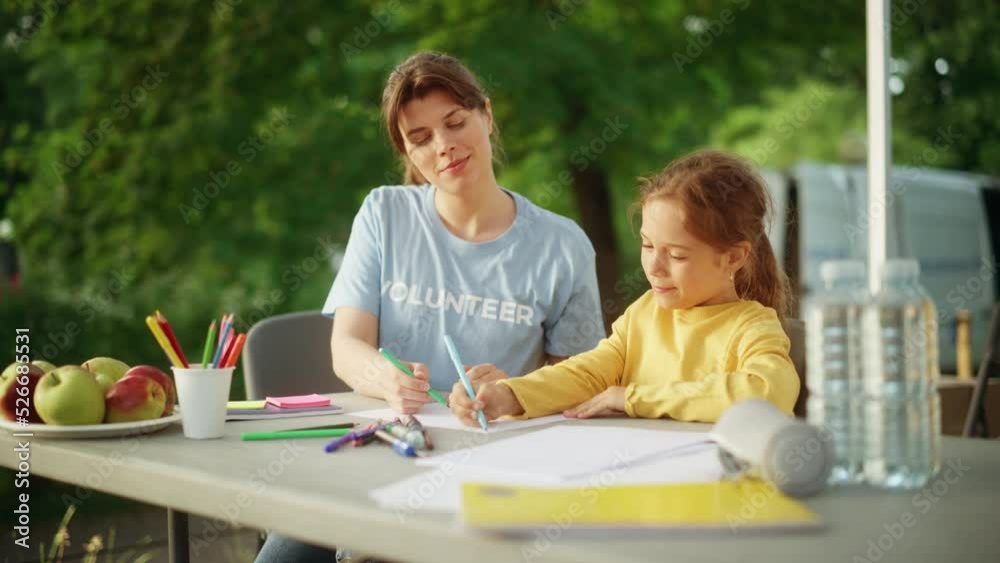 Volunteer Spending Time with a Cute Small Girl, They are Sitting Outdoors and Drawing with Colorful Crayons. Female Charity Worker Teaching a Child to Draw. Helping with Art Homework Assignment. Volunteer Spending Time with a Cute Small Girl, They are Sitting Outdoors and Drawing with Colorful Crayons. Female Charity Worker Teaching a Child to Draw. Helping with Art Homework Assignment.