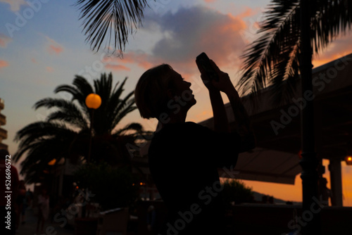 silhouette of a girl taking pictures on her phone against the background of a bright orange sunset and palm leaves. The silhouette of a man at sunset. Gradient of colors.
