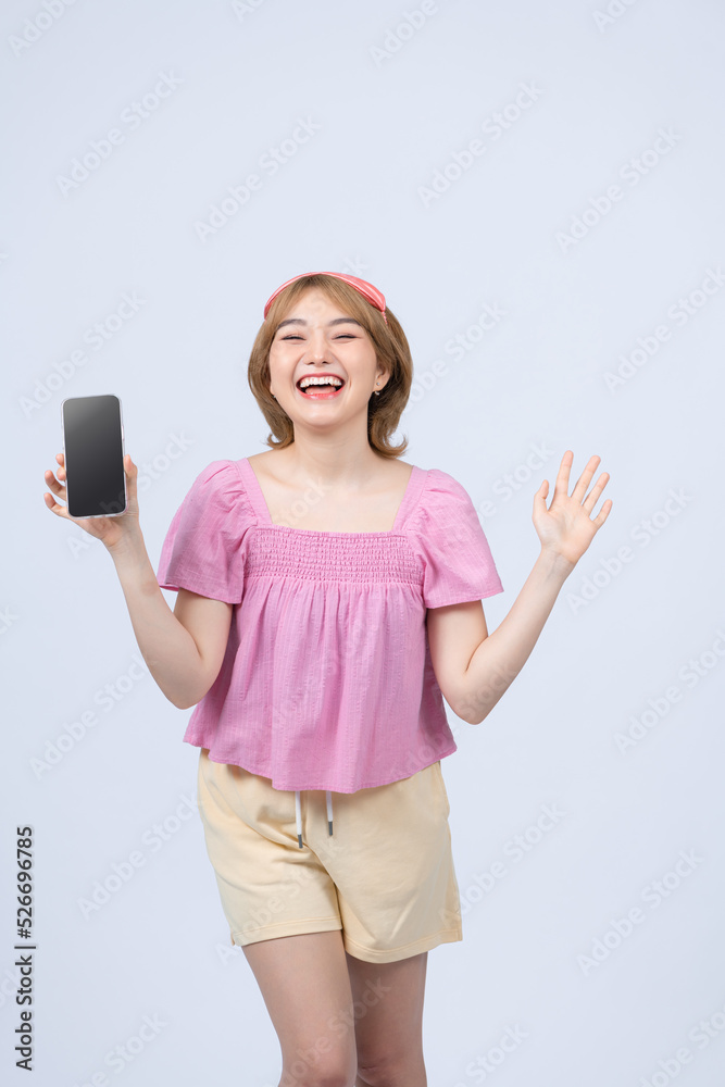 Fototapeta premium Happy smiling asian girl in pajamas and sleeping mask tracking her sleep with mobile app, showing smartphone screen and look pleased, white background