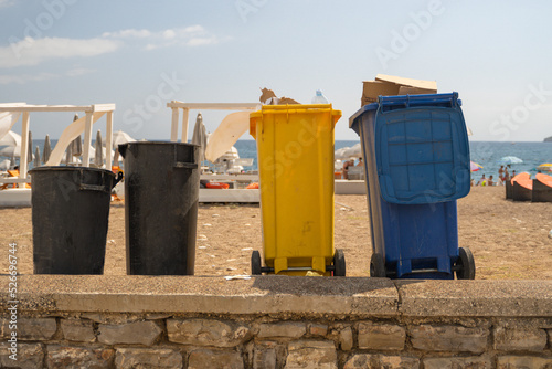colored garbage cans are on the beach for the distribution and sorting of various garbage and waste. Close-up of the garbage recycling system on the beach of the resort town. Blue and yellow trash can © Григорий Галасюк