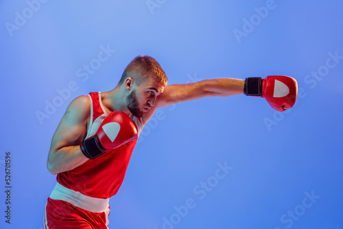 Knockout punch. Male boxer in red uniform and boxing gloves training isolated on blue background in neon. Strength, attack and motion concept. photo