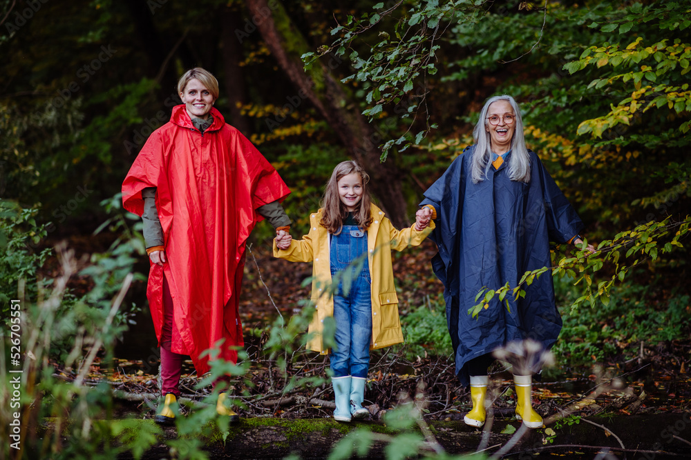 Portrait of small girl with mother and grandmother in raincoats standing by lake outoors in nature.