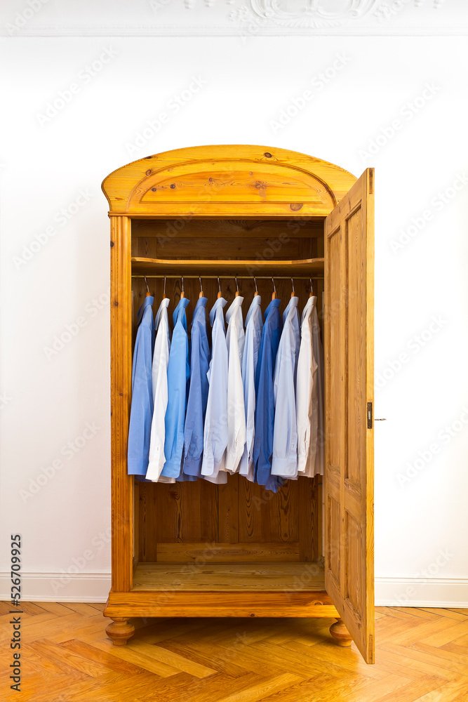 Open antique wardrobe with blue dress shirts, against the white wall of an old building with parquet flooring and stucco plastering, copy space.