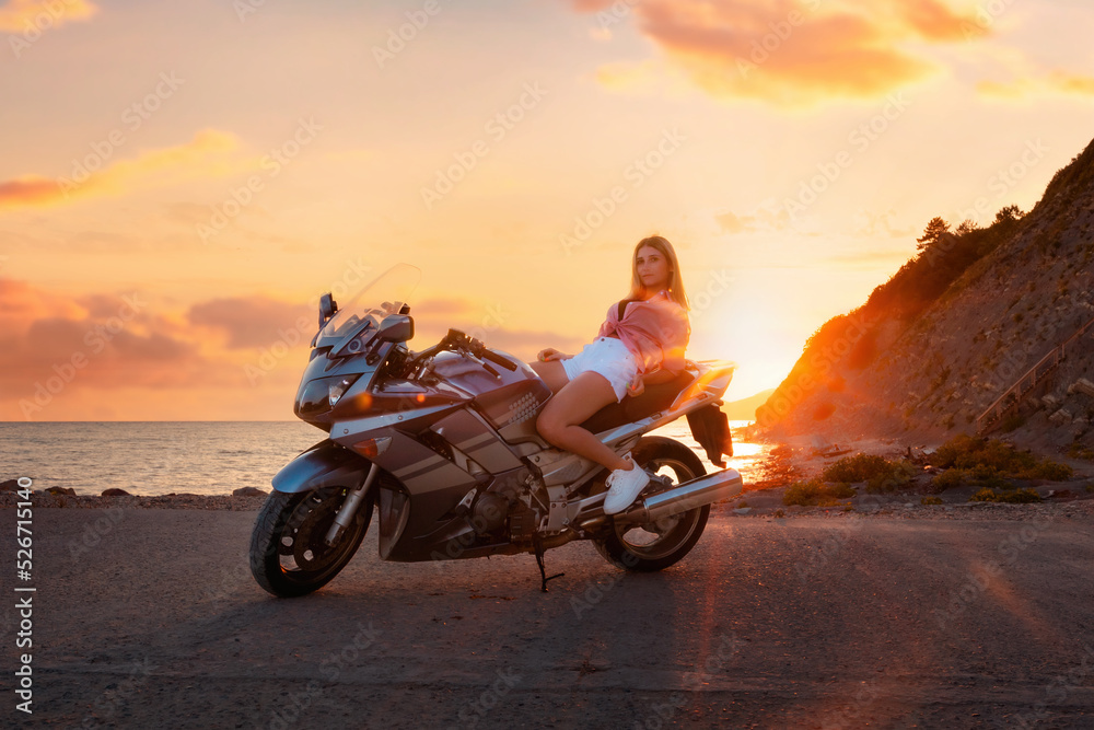 Fototapeta premium World Motorcyclist Day. Caucasian young woman lying posing on a motorcycle. Golden sunset and ocean on the background. Freedom and motorcycle trips