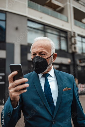 Elegant and positive senior businessman standing on city street and using his smart phone. He is wearing N95 protective face mask as a protection against virus pandemic.