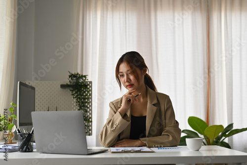 Portrait of a serious Asian businesswoman working on laptop in modern office.