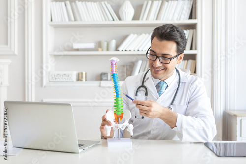 Young handsome male doctor orthopedist demonstrating the problem on spine bone model on the desk in his workplace photo