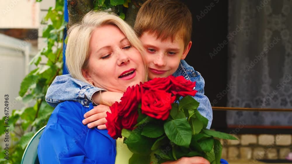 caucasian boy 7-8 years old hugging his blonde mother 40-45 years old sitting outside with a bouquet of red roses.son wishes happy mother's day concept.mom's birthday caucasian boy 7-8 years old hugging his blonde mother 40-45 years old sitting outside with a bouquet of red roses.son wishes happy mother's day concept.mom's birthday