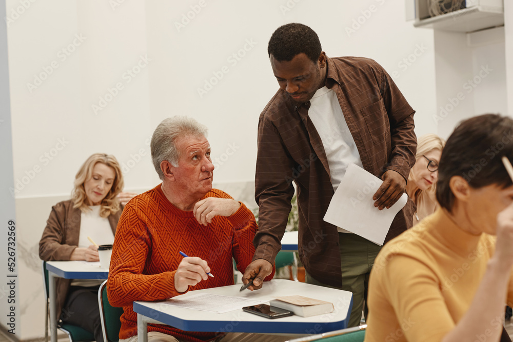 Obraz premium African young teacher walking along the class and explaining adult students how to write test