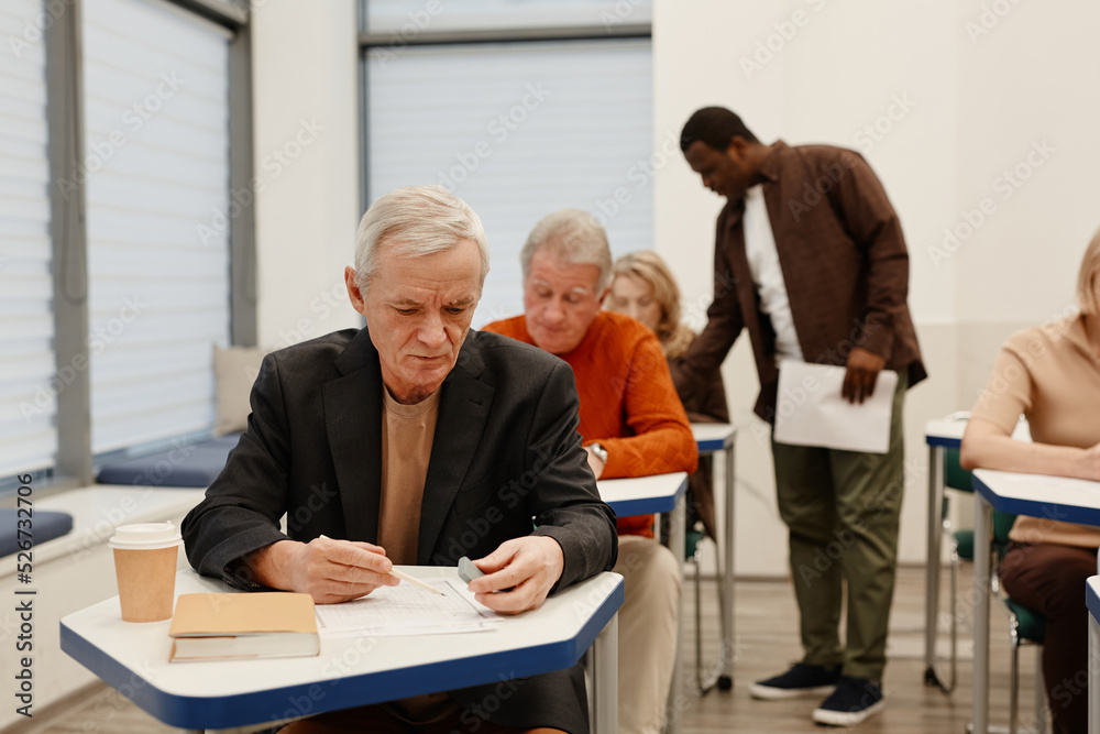Obraz premium Senior students sitting at desks in a row and writing test during lesson while teacher walking along the class