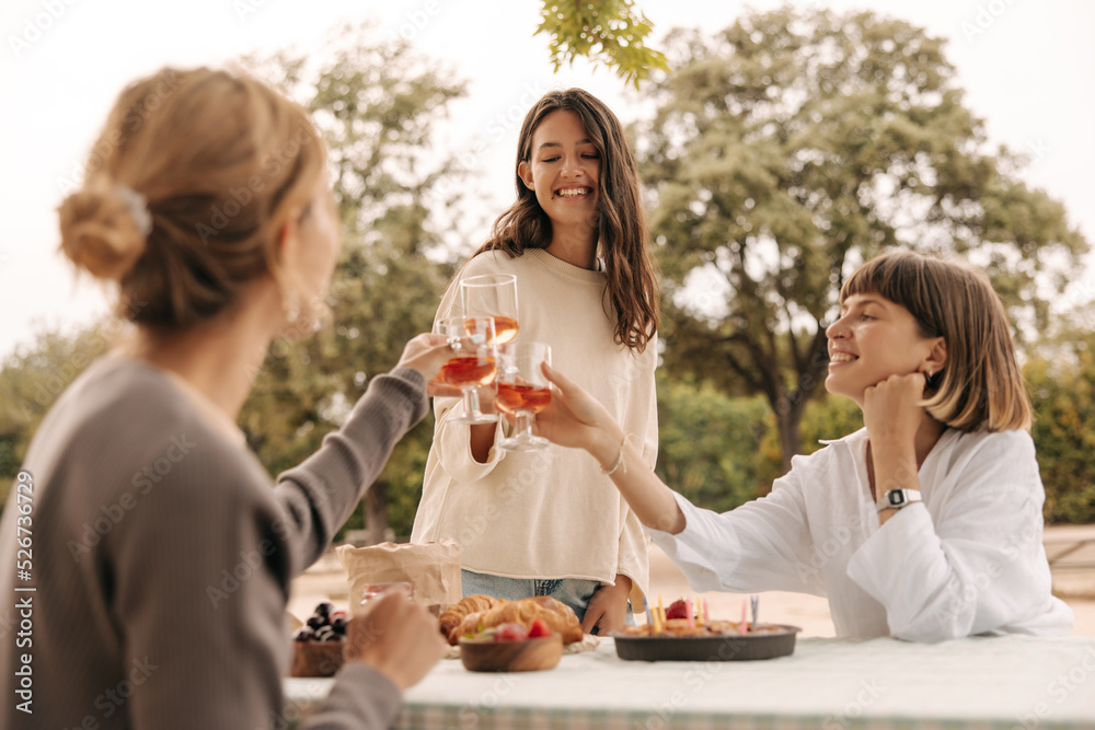 Three nice young caucasian ladies clink glasses, celebrate bachelorette party outdoors spring. Girlfriends wear sweatshirts in warm weather. Rest time concept